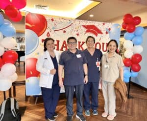 Four people stand in front of a festive "Thank You" backdrop decorated with balloons. Three are wearing scrubs and one is in casual clothing. They appear to be in a celebratory or appreciative setting.