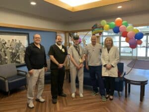 A group of five people in medical attire standing in a hospital lobby. The background includes a framed photo and a colorful balloon decoration.