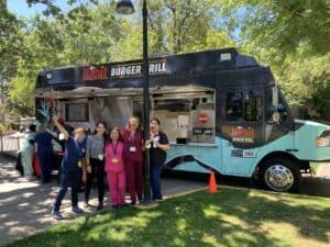 A group of five people in medical scrubs stand in front of a Habit Burger Grill food truck with trees and a lamp post in the background.