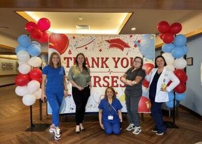Five healthcare professionals are standing in front of a "Thank You Nurses" banner decorated with balloons. Four are standing and one is kneeling, all smiling.