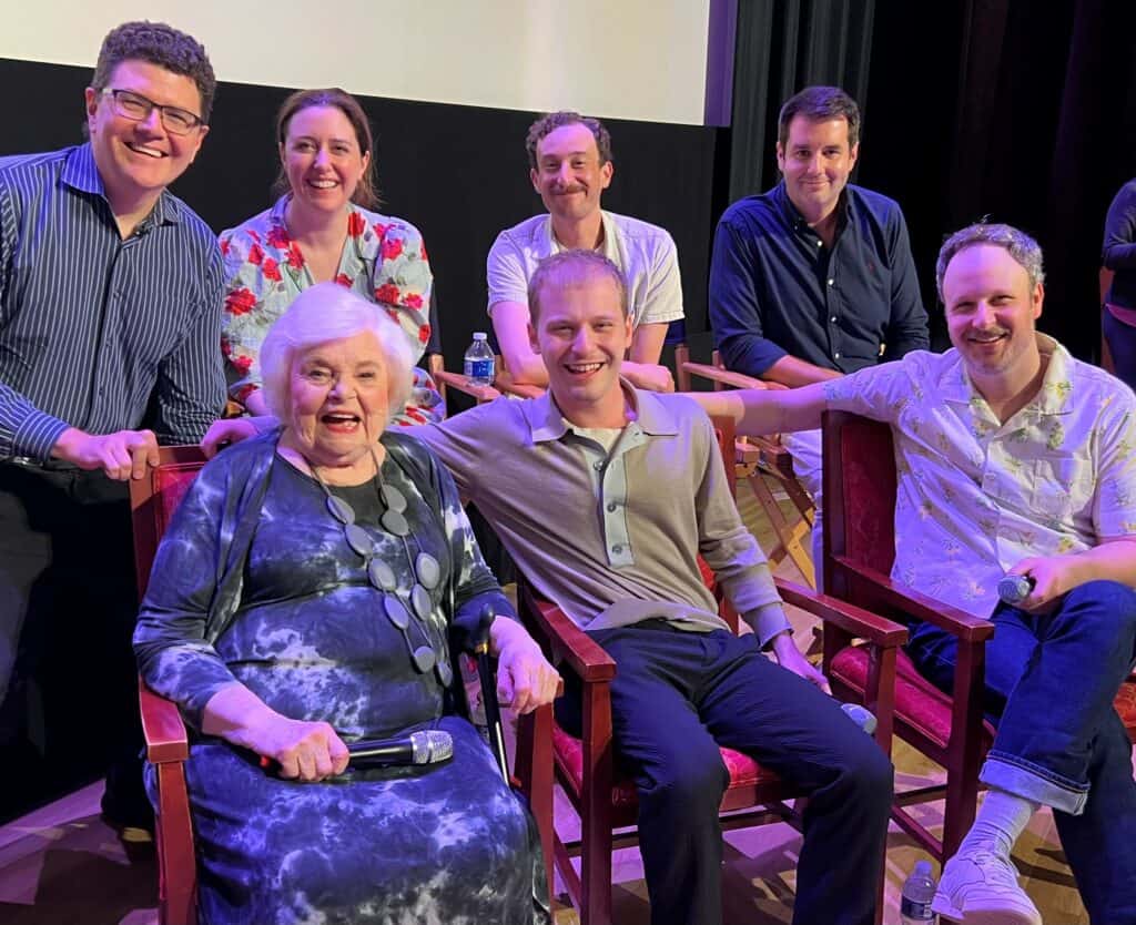 A group of seven people, including an elderly woman holding a microphone, are smiling and posing together in a theater-like setting. Four are seated in chairs, and three are standing behind them.