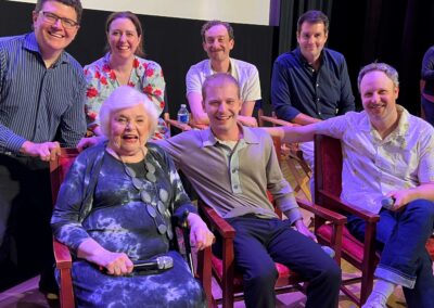A group of seven people, including an elderly woman holding a microphone, are smiling and posing together in a theater-like setting. Four are seated in chairs, and three are standing behind them.