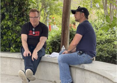 Adam And Tom On Bench Two men sit and talk on a curved concrete bench outdoors, surrounded by greenery. One man holds papers, and a water bottle is placed beside them.